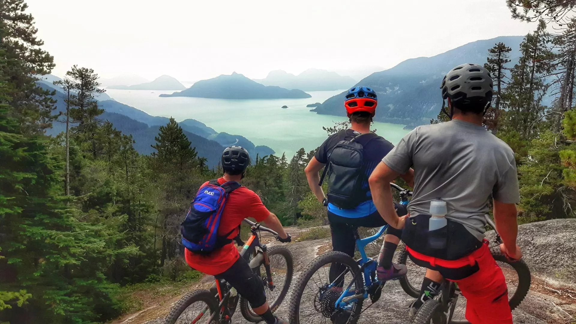 Three adventurous mountain bikers in Squamish, enjoying an adrenaline-pumping ride on a breathtaking trail that offers them panoramic views of a picturesque lake.