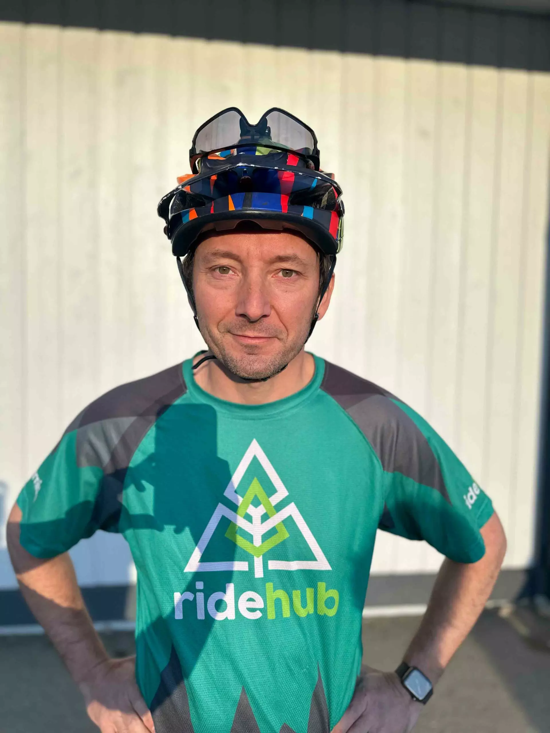 Man in a bicycle helmet and green cycling jersey standing in front of a metal wall, the sun casting shadows on his face.