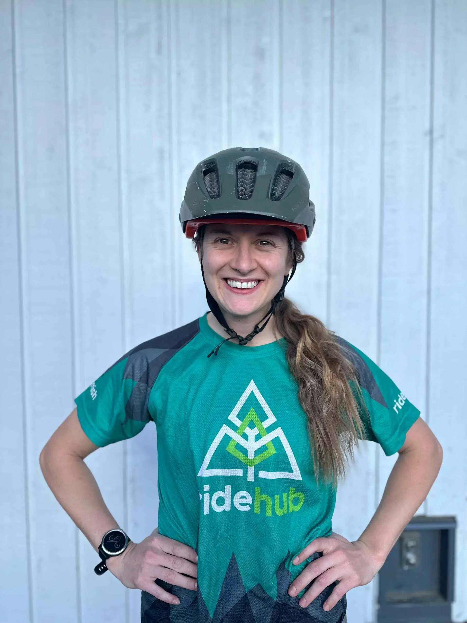 Woman in a bike helmet and blue-green jersey smiling in front of a white corrugated metal background.