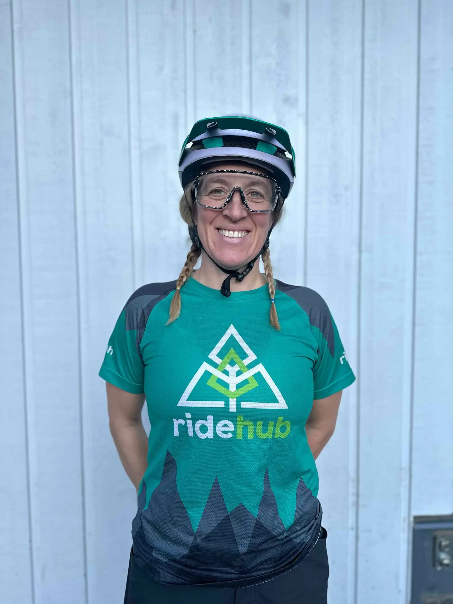 Woman in a green cycling jersey and helmet, smiling at the camera with a corrugated metal background.