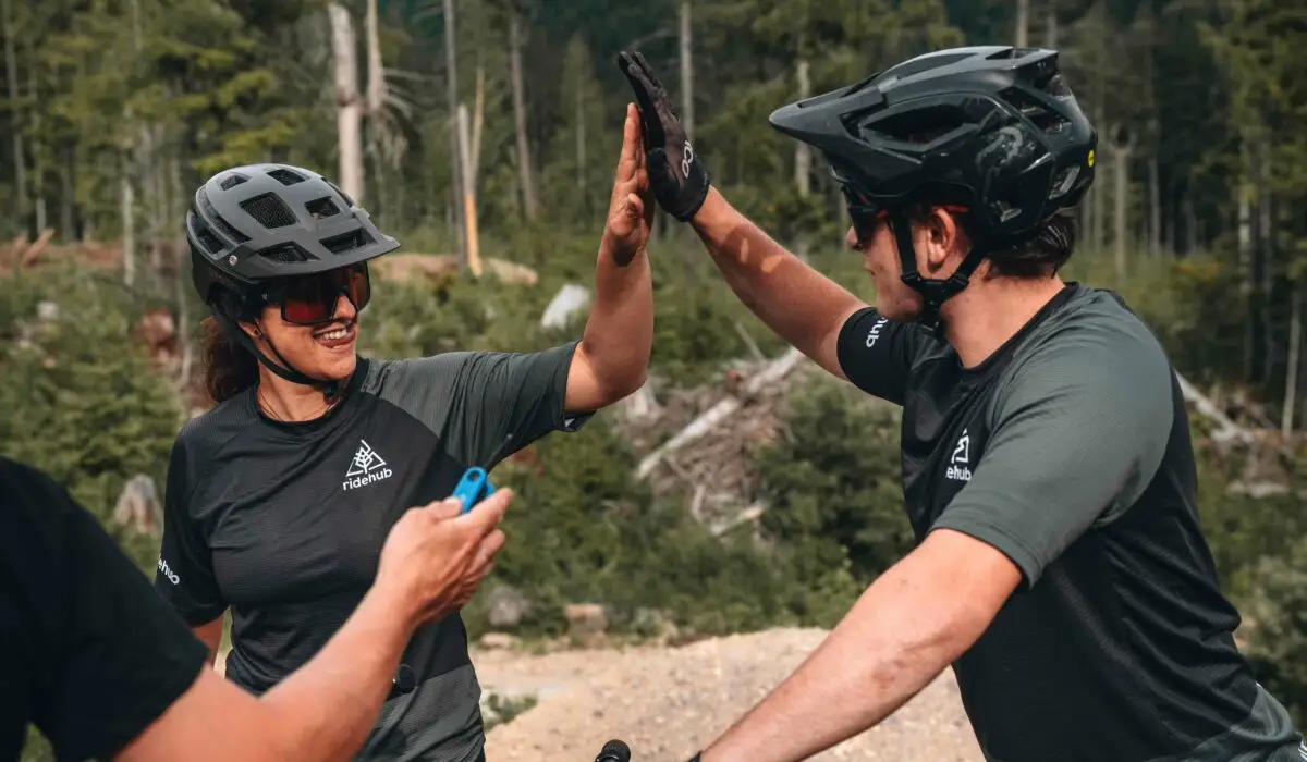 Two cyclists in helmets and sport attire high-five each other in a wooded outdoor setting.