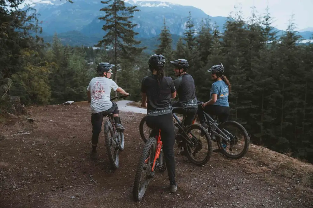 Four cyclists on mountain bikes pause on a trail in a forested area with mountains in the background. They wear helmets and biking gear.