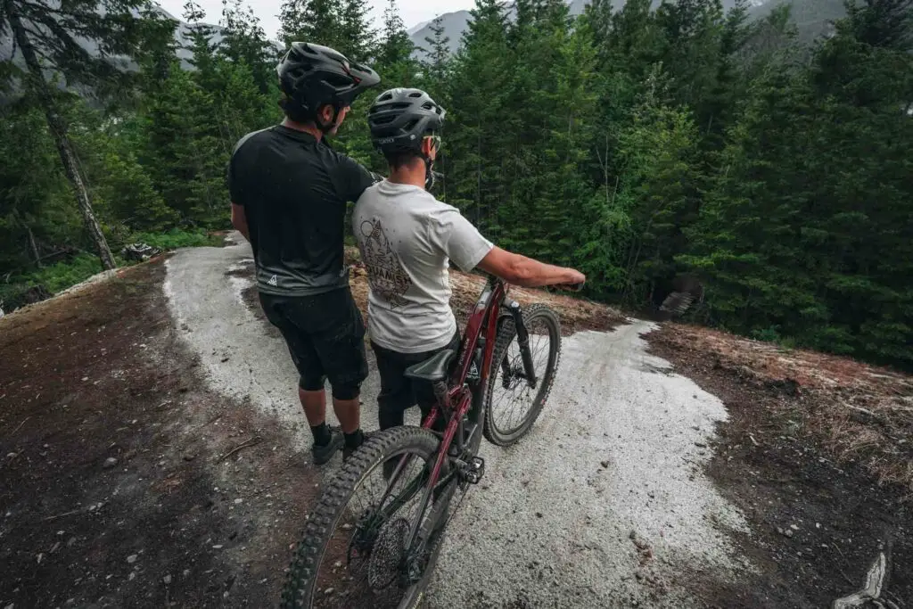 Two cyclists wearing helmets stand on a winding trail in a forest, one with a red mountain bike. They appear to be observing the path ahead.