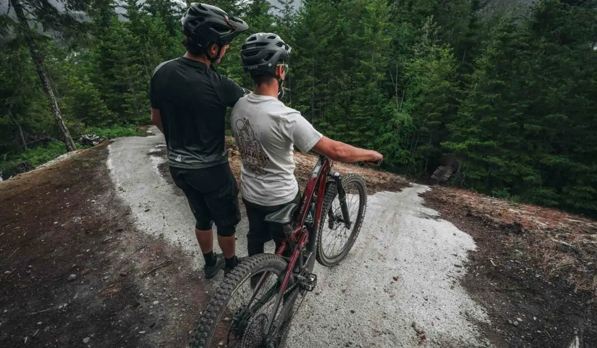 Two cyclists wearing helmets stand on a winding trail in a forest, one with a red mountain bike. They appear to be observing the path ahead.