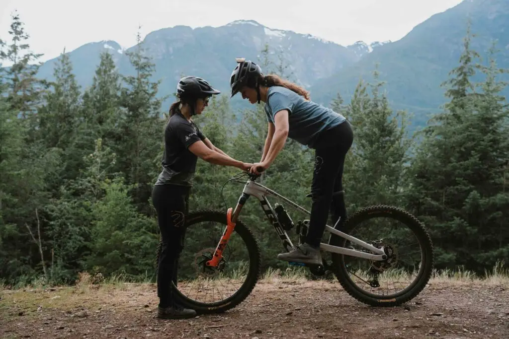 Two people with helmets stand in a wooded area with mountains in the background; one person stands beside a bicycle while the other, on the bike, appears to be giving instructions or assistance.