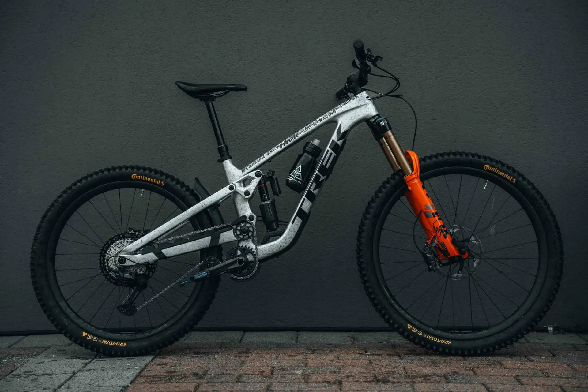 A gray Trek mountain bike with orange front suspension and black tires is positioned against a dark gray wall.