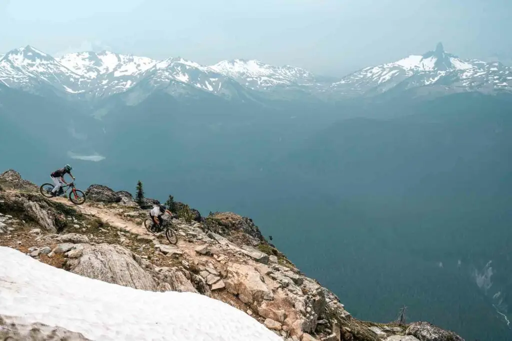 Two cyclists ride on a mountain trail with snow patches, with a background of snow-capped peaks and a wide valley below.