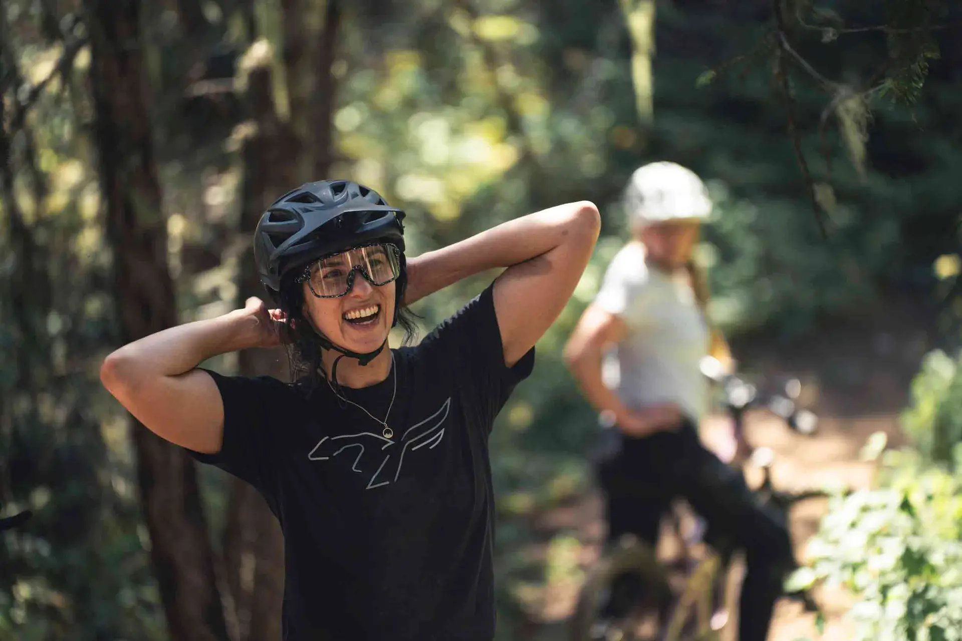 A person in a black helmet and glasses smiles while putting their hands behind their head. Another person in a white helmet stands with a bike in the background on a forest trail.