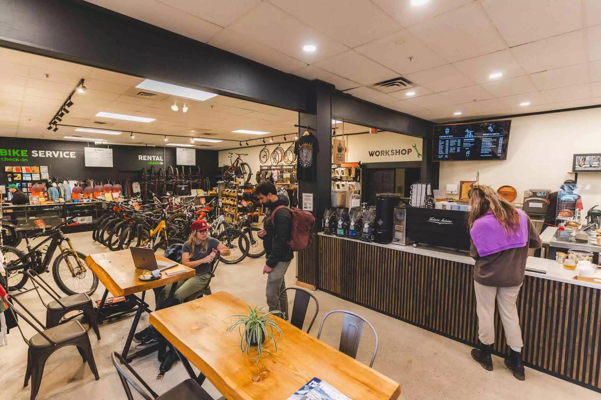 A bike shop and café bustling with customers and employees. Mountain bikes are displayed on the left, a counter with a menu board on the right, and people interacting or working on laptops while a mechanic expertly tunes bikes in the background.