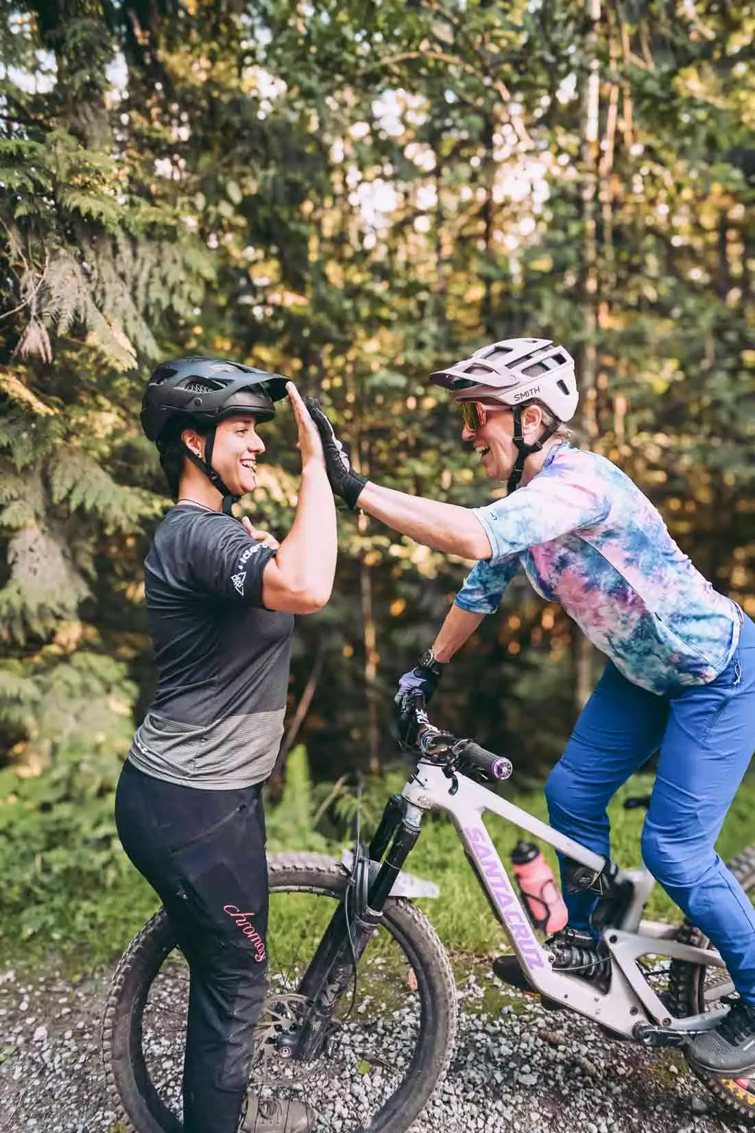 Two people in helmets high-five near a mountain bike on a forest trail, celebrating their adventurous ride through the picturesque paths of British Columbia.