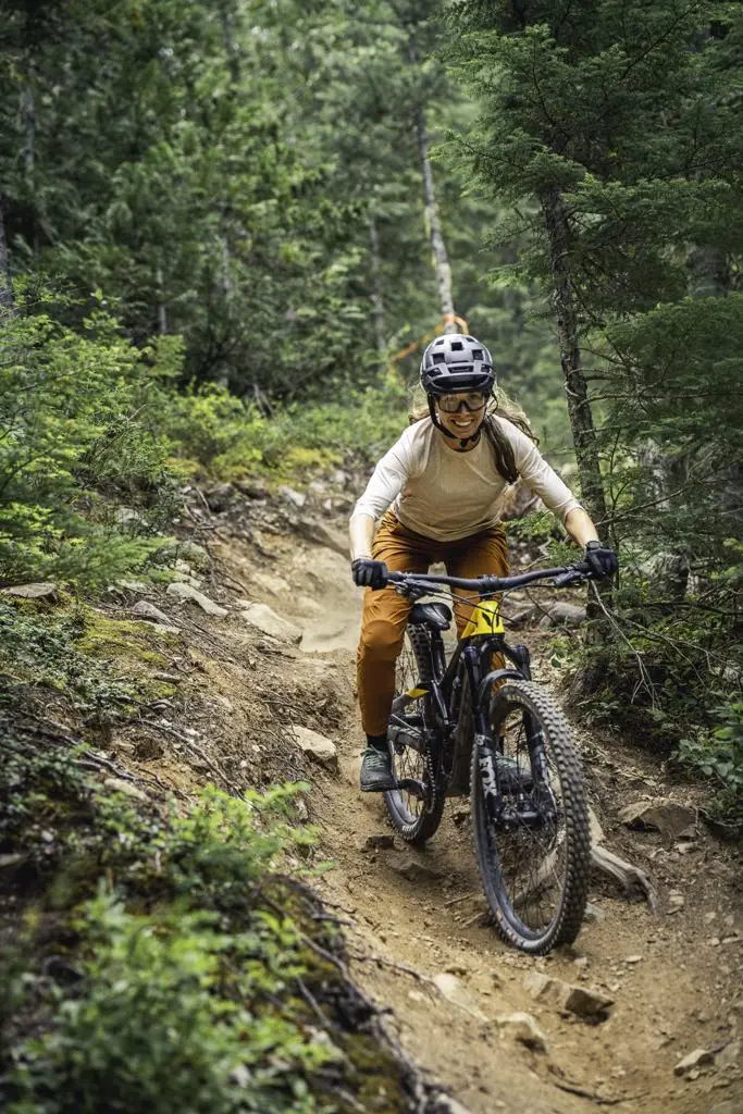 A person wearing a helmet and gloves rides a mountain bike down a narrow, rocky forest trail surrounded by greenery.